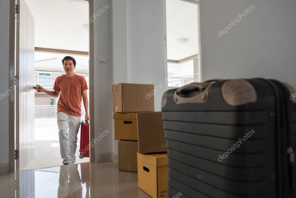 Portrait of Asian husband man open front door and carry luggage to new house with many moving cardboard boxes inside empty home. Family start new life. Real estate buy or purchase concept.