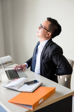 Resmi üniformalı Asyalı iş adamı ofisteki çalışma masasında acı dolu duygularla arkasını kolluyor. İşletme patronu ofis sendromu yaşar. Sağlık, sigorta, sıkı çalışma konsepti.