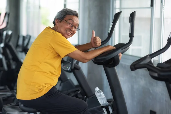 Happy senior aged man with gray hair exercising on cycling machine and thumb up in fitness gym. Old mature fit people biking cardio. Bodybuilding and elderly Healthy lifestyle.