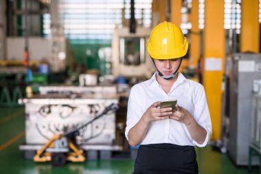 Miğferli kadın işçi akıllı telefondaki şirket uygulaması ile fabrika deposunda üretim malzemelerini kontrol ediyor. Sanayi ve teknoloji üretimi. Envanter konsepti olan endüstri.