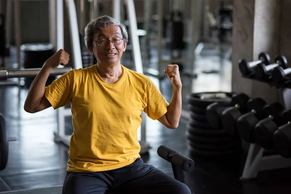 Old man show bicep muscle after dumbbell exercise alone in fitness gym. Bodybuilding and healthy lifestyle during covid-19 pandemic disease. Quarantine, stay home, wfh or work home, social distancing.