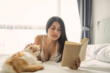 Pijamalı seksi Asyalı kadın kitap okuyor ve beyaz yatakta sarı egzotik bir kediyle oynuyor. Covid-19 salgını sırasında evde kalmak. WFH ya da evden çalışma, karantina, sosyal uzaklık.