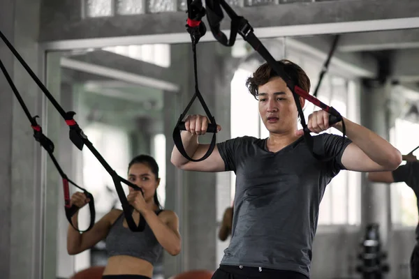 American sweat man and Asian woman couple exercise with abdominal straps at fitness gym. Bodybuilding sport and healthy lifesytle for happy couple.