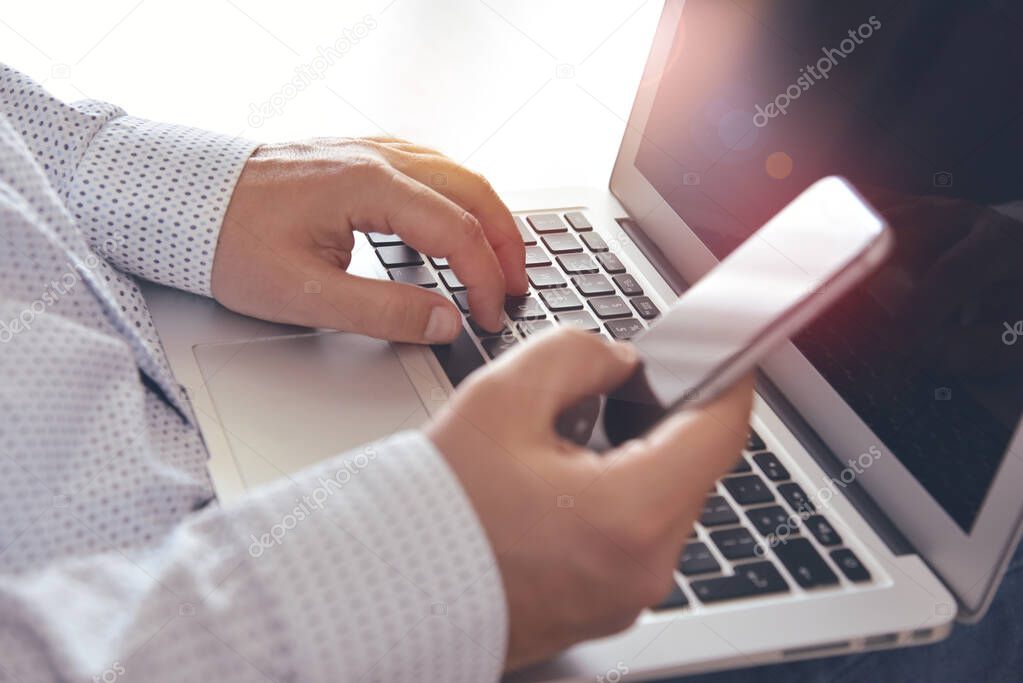 Closeup of man's hands holding mobile phone and typing on laptop keyboard. Concept for modern online communications, e-commerce, internet services and apps.