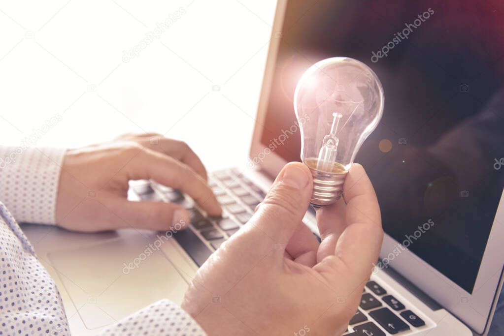 Smart idea. Man's hands holding electric bulb and typing on laptop keyboard.