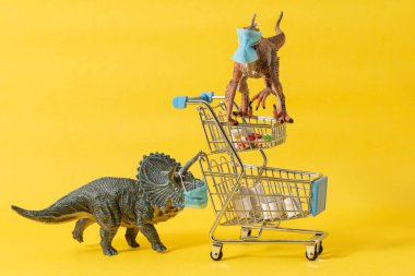 Yüz maskeli iki plastik dinozor ve sarı arka planda bakkaliye arabaları.