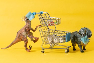 Yüz maskeli iki plastik dinozor ve sarı arka planda bakkaliye arabaları.