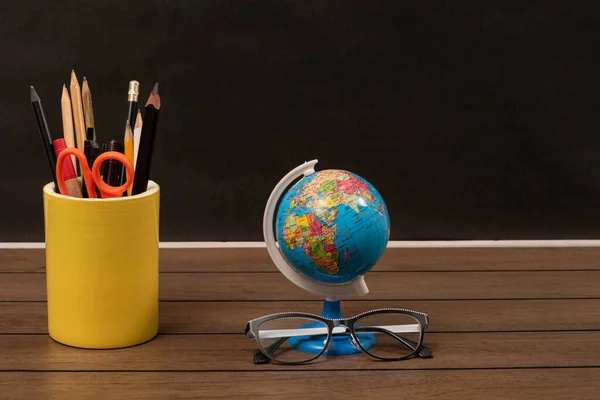 A stand with pencils and scissors, a globe and glasses on a wooden table on black background