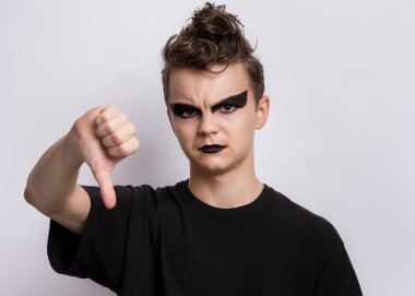 Ürkütücü makyajlı genç çocuk olumsuz ifade ve hoşnutsuzlukla başparmak hareketi yapıyor. Siyahlar giymiş punk tarzı gotik bir genç, kötü sinyal veriyor. Gri arkaplandaki çocuk.