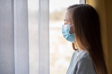 Koronavirüs karantinası kavramı. Grip salgını sırasında koruyucu yüz maskesi takan çocuk pencereden dışarı bakıyor. COVID-19, kendini izole etme. Genç kız evde kalmaya zorlandı. Önleme salgını.
