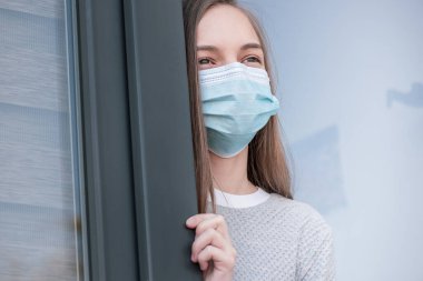 Koronavirüs karantinası kavramı. Grip salgını sırasında koruyucu yüz maskesi takan çocuk pencereden dışarı bakıyor. COVID-19, kendini izole etme. Genç kız evde kalmaya zorlandı. Sokaktan görüntüle.