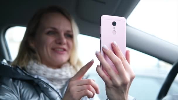 Blogueira loira desfocada em roupas de inverno sentada no carro e conversando com seguidores, transmissão ao vivo, olhando para a tela do smartphone. foco no telefone — Vídeo de Stock