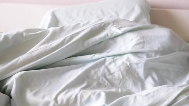 Um menino loiro deita-se na cama e sorri, está se divertindo, escondendo-se sob os lençóis e espreitando debaixo dele no início da manhã. Hora de acordar — Vídeo de Stock