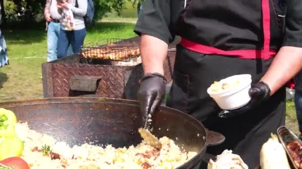 Siyah elbiseli ve eldivenli bir aşçı, büyük bir kazanın sokak yemeklerinin olduğu tabağa güzel kokulu pilav koyar. Sokak festivali taze hazırlanmış yiyecekler. Et ve pilav. Geleneksel Asya mutfağı.. — Stok video