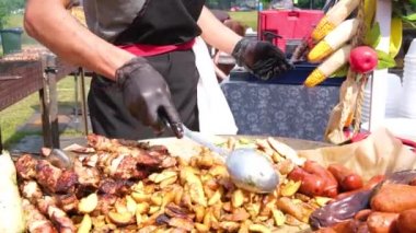 Sokak Yemekleri Festivali. Siyah elbiseli ve eldivenli bir şef yeni bir yığın patates ekler. Patates, ızgara sosis, çubuklu et, barbekü. Yemek yakın.