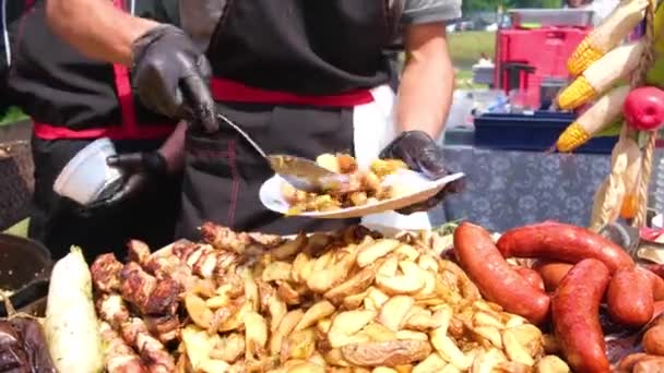 Street Food Festival. En kock i svarta kläder och handskar sprider stekt mat på tallrikar. Potatis, grillad korv, kött med pinnar, grill. Närbild av livsmedel. — Stockvideo