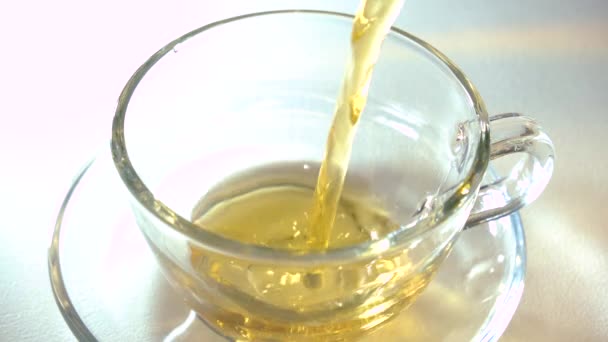 Pouring aromating black tea from teapot into a matching transparent glass tea cup on white background closeup. — Stock Video