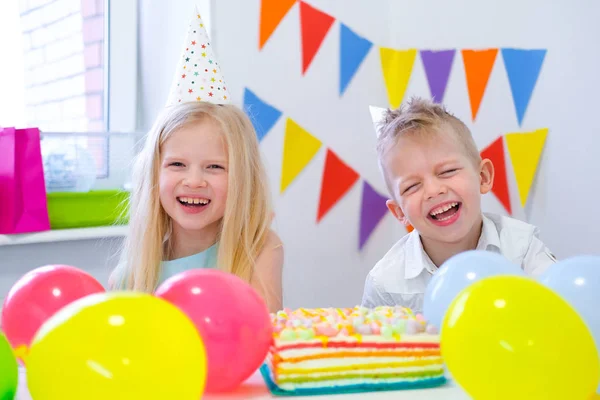 Dos niños caucásicos rubios chico y chica divirtiéndose y riendo en la fiesta de cumpleaños. Fondo colorido con globos y pastel de arco iris de cumpleaños . —  Fotos de Stock