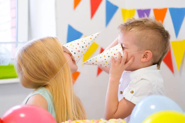 Dos niños caucásicos rubios chico y chica divirtiéndose jugando con sombreros en la fiesta de cumpleaños. Fondo colorido con globos —  Fotos de Stock