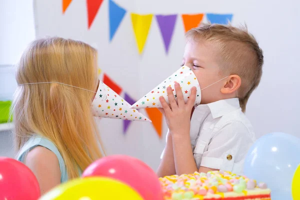 Duas crianças loira caucasiana menino e menina se divertindo brincando com chapéus na festa de aniversário. Fundo colorido com balões — Fotografia de Stock