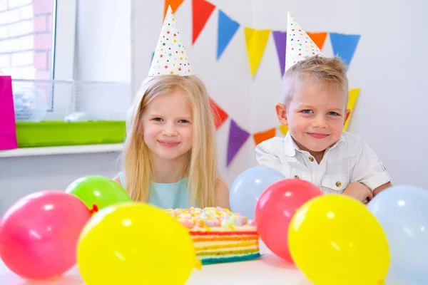 Duas crianças loira caucasiana menino e menina em chapéus de aniversário olhando para a câmera e sorrindo na festa de aniversário. Fundo colorido com balões e bolo de aniversário arco-íris . — Fotografia de Stock