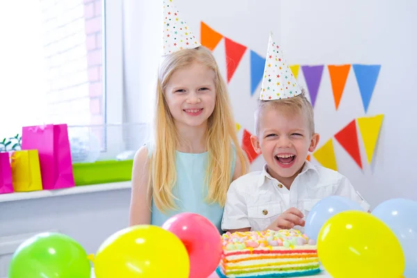 Dos niños caucásicos rubios niño y niña en sombreros de cumpleaños mirando a la cámara y sonriendo en la fiesta de cumpleaños. Fondo colorido con globos y pastel de arco iris de cumpleaños . —  Fotos de Stock