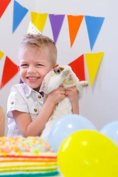 Feliz chico caucásico rubia abrazando a su conejo presente y riendo en la fiesta de cumpleaños. Fondo colorido con globos y pastel de arco iris de cumpleaños . — Foto de Stock