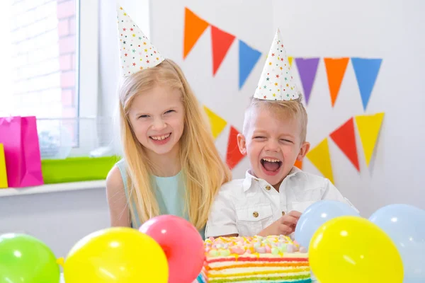 Two blonde caucasian kids boy and girl having fun and laughing at birthday party. Colorful background with balloons and birthday rainbow cake. Stock Picture