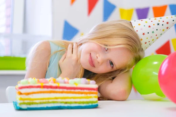 Chica rubia caucásica sonriendo a la cámara cerca de pastel de arco iris de cumpleaños. Fondo colorido festivo con globos —  Fotos de Stock