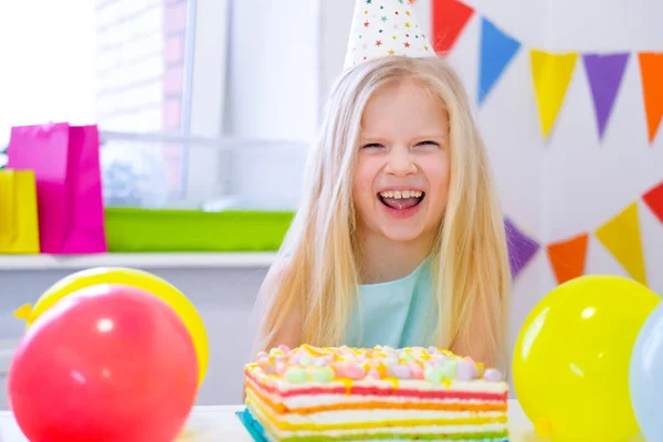 Blonde caucasian girl laughing at camera near birthday rainbow cake. Festive colorful background with balloons — 스톡 사진