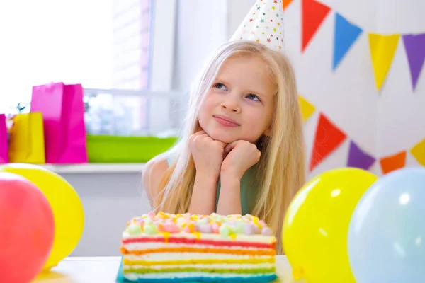 Chica rubia caucásica se sienta pensativa y soñadoramente en la mesa festiva cerca de pastel de arco iris de cumpleaños y pide un deseo. Fondo colorido con globos —  Fotos de Stock