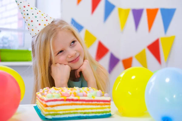 Chica rubia caucásica se sienta pensativa y soñadoramente en la mesa festiva cerca de pastel de arco iris de cumpleaños y pide un deseo. Fondo colorido con globos — Foto de Stock