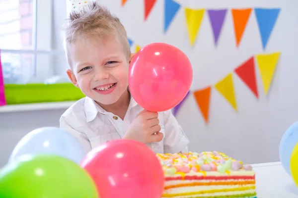 Ritratto di ragazzo caucasico biondo che sorride alla telecamera vicino alla torta arcobaleno di compleanno. Sfondo colorato festivo con palloncini — Foto Stock