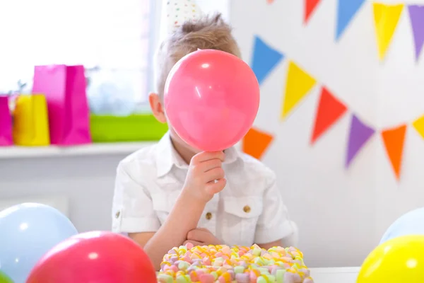 Blond kaukasisk pojke gömde sig bakom en röd ballong nära födelsedag regnbåge tårta. Festlig färgglad bakgrund. Rolig födelsedagsfest — Stockfoto