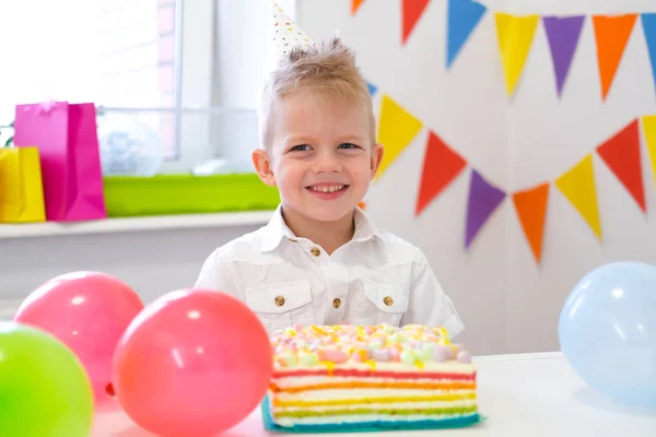 Portrait of blonde caucasian boy smiling at camera near birthday rainbow cake. Festive colorful background with balloons — 스톡 사진