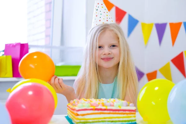 Sarışın beyaz kız doğum günü pastasının yanında kameraya gülümsüyor. Balonlarla şenlikli bir arkaplan Stok Fotoğraf