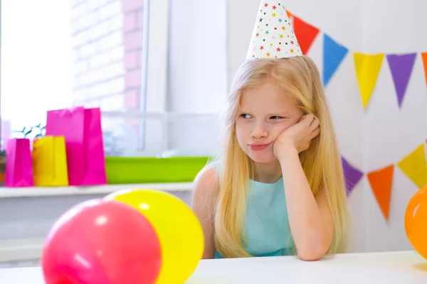 Chica caucásica rubia infeliz con cara aburrida cerca de pastel de arco iris de cumpleaños. Fondo colorido festivo. Mala fiesta de cumpleaños —  Fotos de Stock