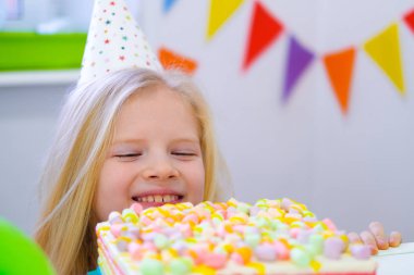 Doğum günü partisinde gülümseyen yüzüyle doğum günü pastasının arkasından dışarı bakan beyaz sarışın kız. Balonlarla şenlikli bir arkaplan.