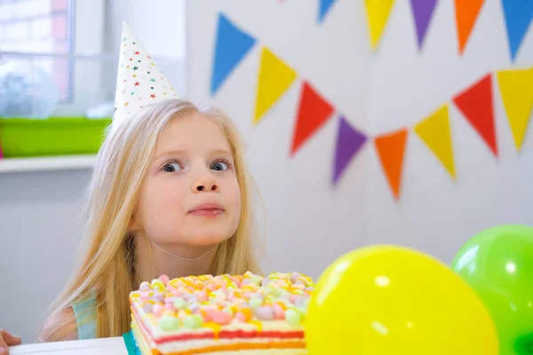 Menina loira caucasiana espreitando por trás bolo de aniversário com um rosto engraçado na festa de aniversário. Fundo colorido festivo com balões . — Fotografia de Stock