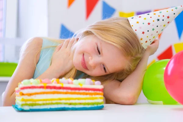 Blonde caucasian girl dreamily smiling near birthday rainbow cake. Festive colorful background with balloons — 스톡 사진
