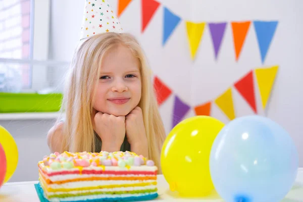 Chica rubia caucásica se sienta pensativa y soñadoramente en la mesa festiva cerca de pastel de arco iris de cumpleaños y pide un deseo. mirando a la cámara. Fondo colorido con globos —  Fotos de Stock