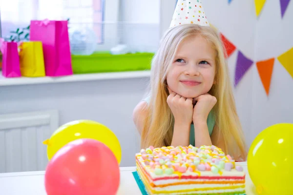 Chica rubia caucásica se sienta pensativa y soñadoramente en la mesa festiva cerca de pastel de arco iris de cumpleaños y pide un deseo. Fondo colorido con globos —  Fotos de Stock