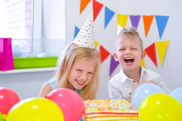 Dos niños caucásicos rubios chico y chica divirtiéndose y riendo en la fiesta de cumpleaños. Fondo colorido con globos y pastel de arco iris de cumpleaños . —  Fotos de Stock