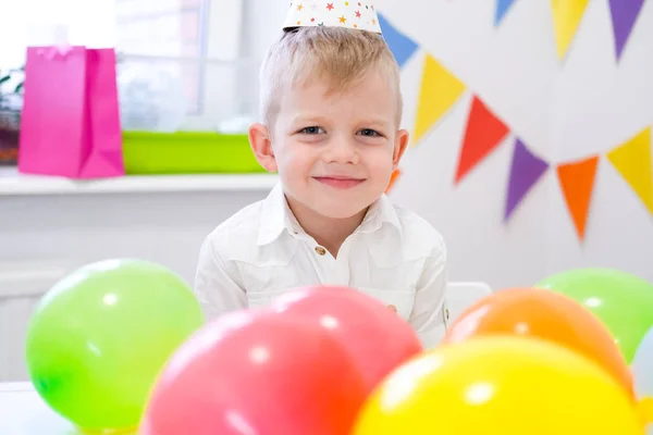 Portrait of blonde caucasian boy smiling at camera near birthday rainbow cake. Festive colorful background with balloons — 스톡 사진