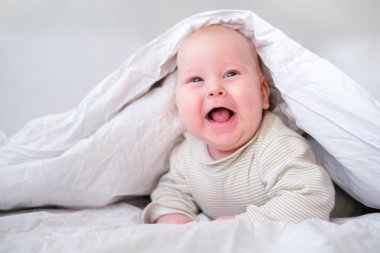 Yatak odasında beyaz battaniyenin altında gülümseyen sevimli bir oğlan. Yeni doğmuş çocuk yatakta dinleniyor. Aile sabahı evde. Yeni doğmuş bebekler.
