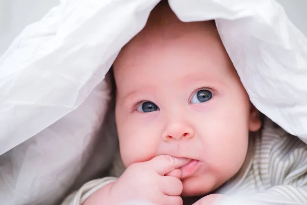 El bebé infantil pensativo yace debajo de una manta blanca en la cama y sostiene el dedo en su boca. Niño recién nacido relajándose en la cama. Concepto de niños recién nacidos — Foto de Stock