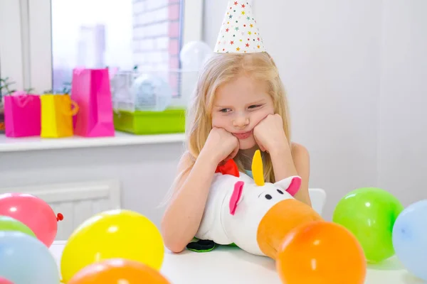 Chica caucásica rubia infeliz con cara aburrida cerca de pastel de arco iris de cumpleaños. Fondo colorido festivo. Mala fiesta de cumpleaños — Foto de Stock