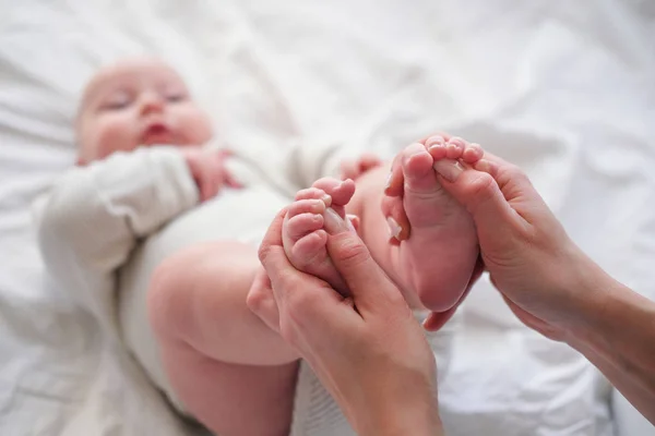 Pieds de bébé entre les mains de la mère. Jeune femme caucasienne fait massage pour bébé heureux sur lit blanc à la maison. Garde d'enfants, sport et maternité heureuse . — Photo