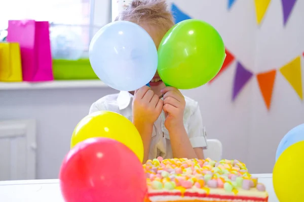 Cumpleaños niño se escondió detrás de globos de colores cerca de pastel de arco iris de cumpleaños. Fondo del festival. Fiesta de cumpleaños divertida —  Fotos de Stock