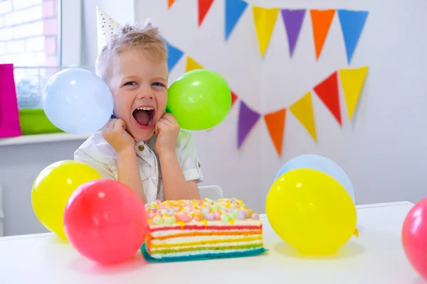 Garoto de aniversário animado com balões coloridos perto de bolo de aniversário arco-íris. Fundo festivel. Festa de aniversário engraçado — Fotografia de Stock